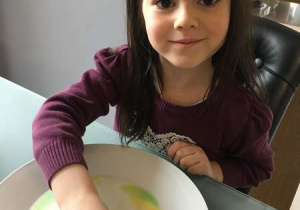 Dziewczynka trzyma patyczek kosmetyczny w mleku z barwnikami na talerzyku – wykonuje doświadczenie łączenia się kolorów.